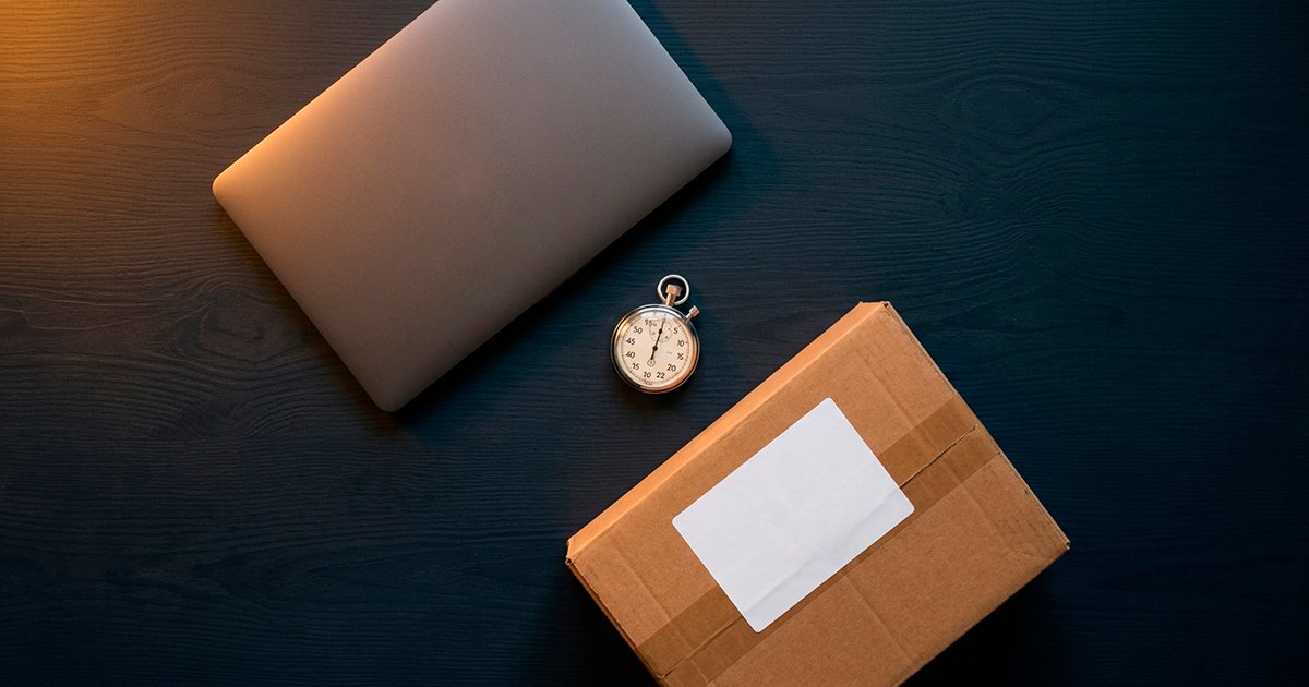 Portátil cerrado junto a un cronómetro y una caja de envío sobre un escritorio oscuro, representando la rapidez del proceso para vender un computador usado